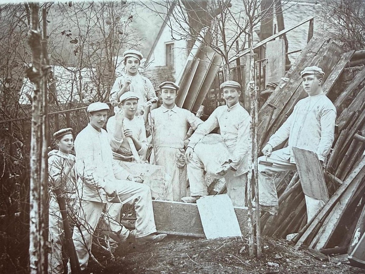 Historische Schwarzweißaufnahme: eine Gruppe von Handwerkern in heller Arbeitskleidung mit Werkzeug, vor Brettern und einem Wohnhaus.