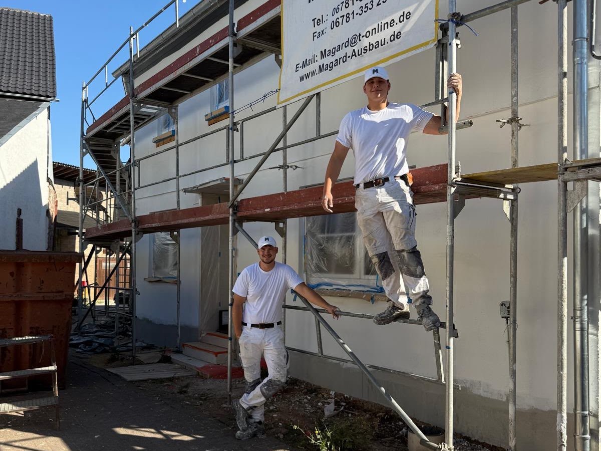 Zwei junge Handwerker in weißer Arbeitskleidung am Gerüst eines Wohnhauses, darüber das Firmenschild des Betriebs.