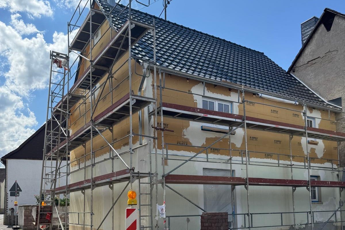 Wohnhaus im Gerüst: auf der Fassade sind Dämmplatten angebracht, daneben liegt der alte Putz noch frei. Am Gehweg steht eine Absperrbake.