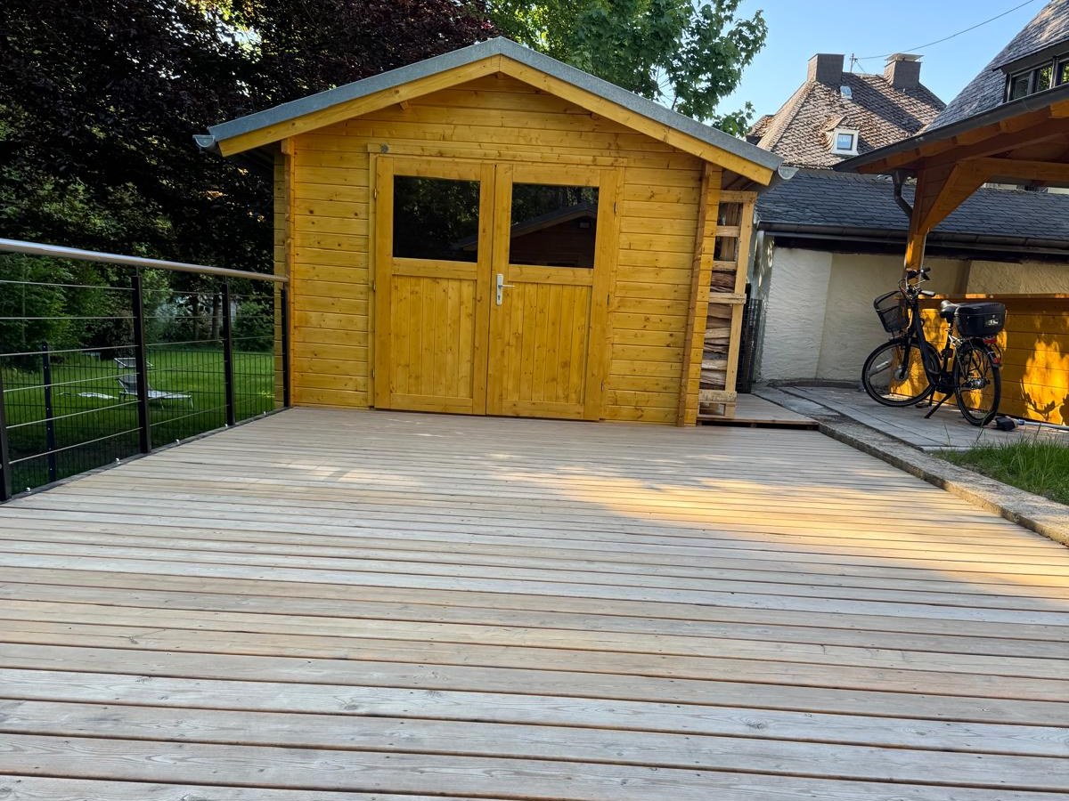 Holzterrasse mit unbehandelten, hellgrauen Dielen vor einem Gartenhaus.