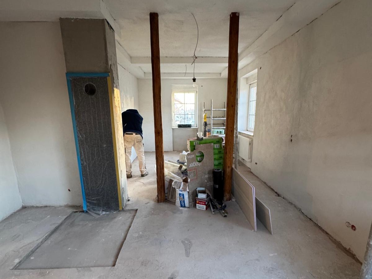 Raum im Ausbau mit frisch verputzten Wänden, freistehenden Holzstützen und Baumaterial auf dem Estrich.