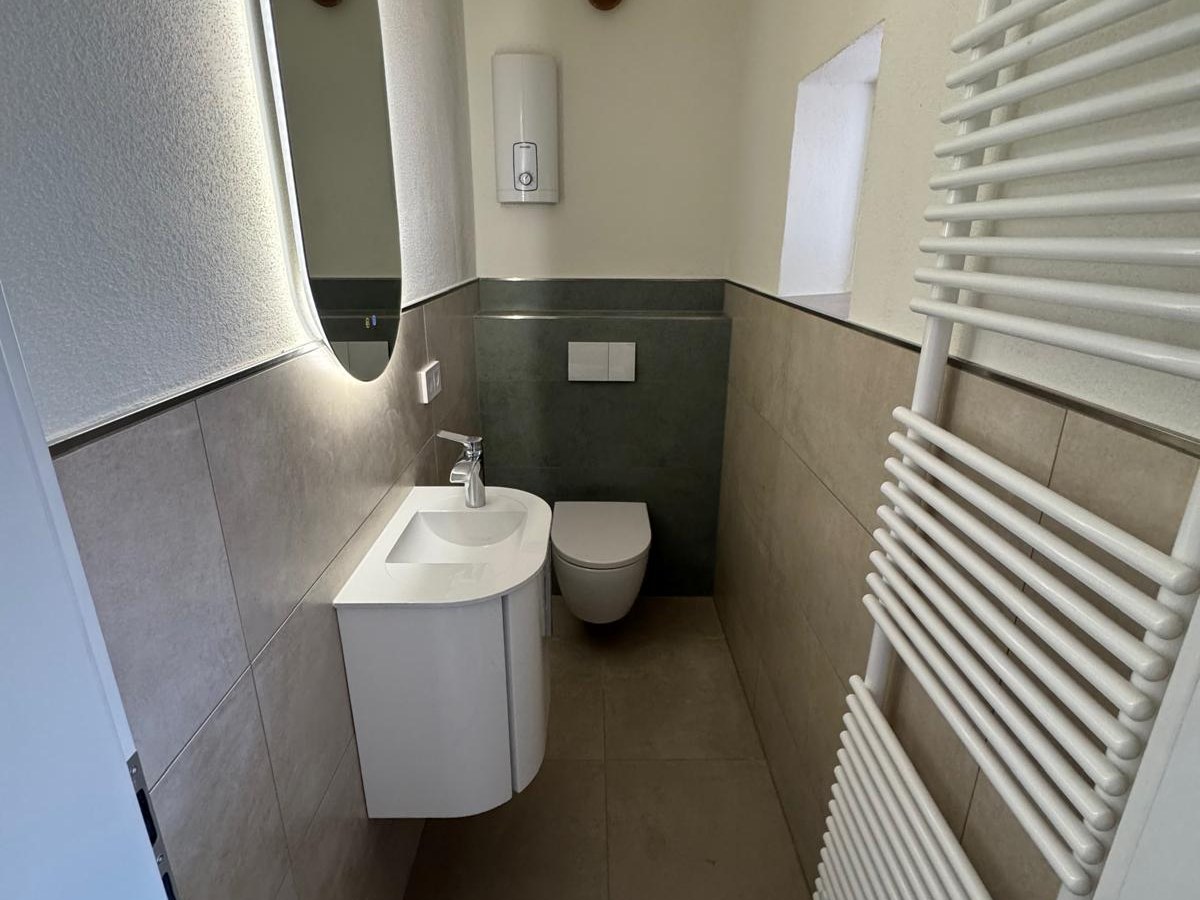 Dasselbe Gäste-WC fertig gefliest, mit Waschtisch, beleuchtetem Spiegel und Handtuchheizkörper.